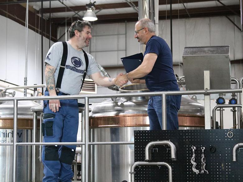 An instructor and a student shake hands in front of some brew kettles